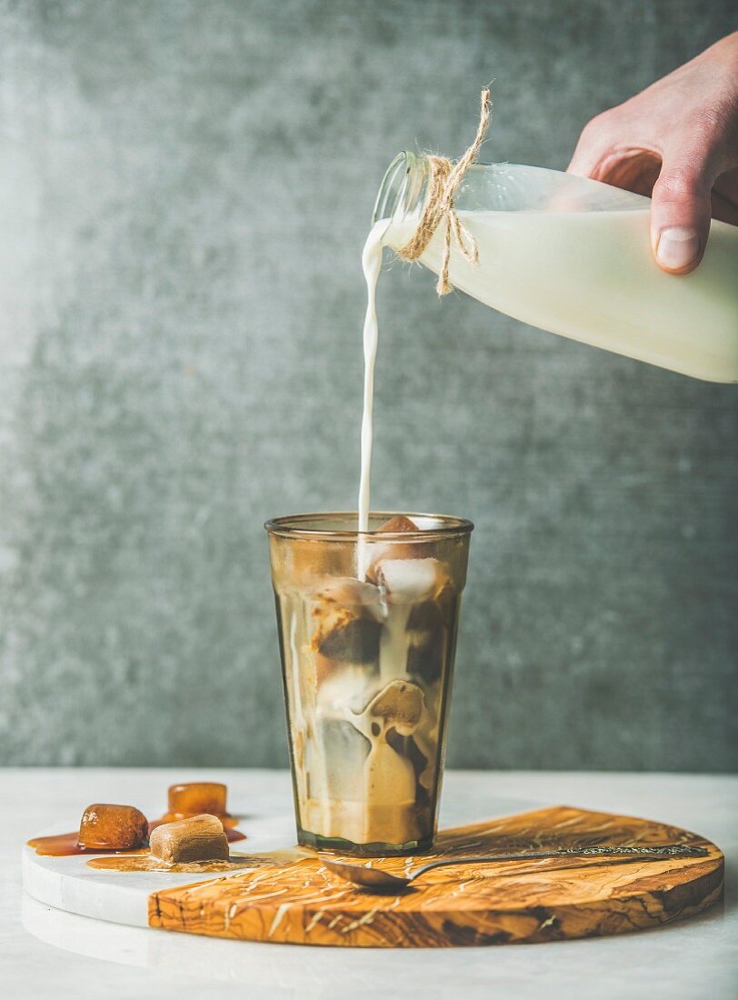Hand giesst Milch aus der Flasche in Glas mit Kaffee- und Karamelleiswürfeln