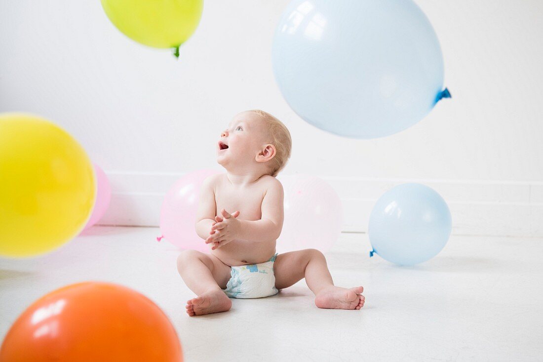 A baby boy wearing a nappy sitting on the floor and playing with balloons