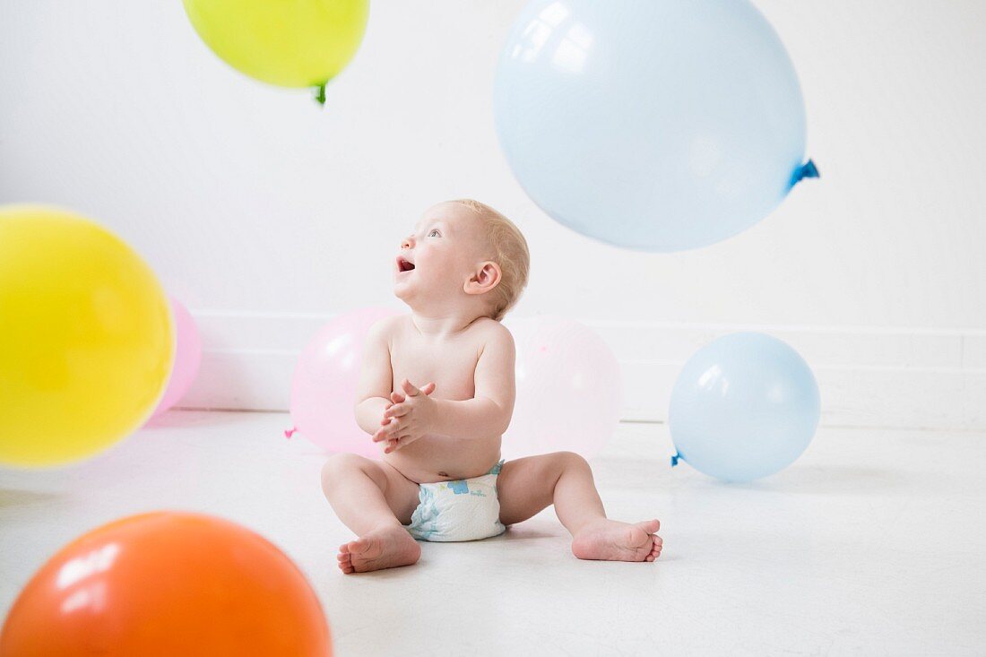 Baby sitzt in Pampers auf dem Boden und spielt mit Luftballons