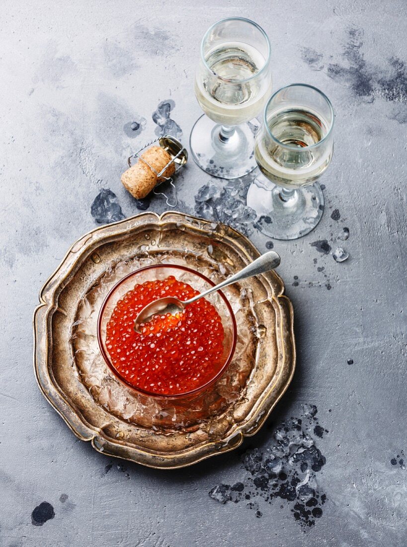 Red caviar in glass bowl on ice and champagne on concrete background