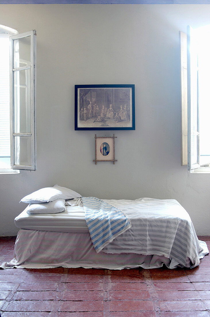 Bed covered in throw on terracotta-tiled floor between two windows