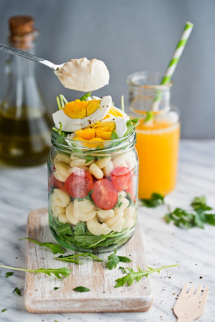 A spoonful of mayonnaise being held over pasta salad with tomatoes, egg and rocket in a glass jar