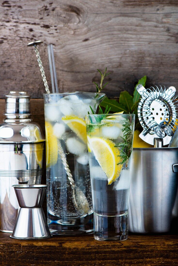 Gin and tonic with lemon, ice cubes and rosemary between various bar utensils