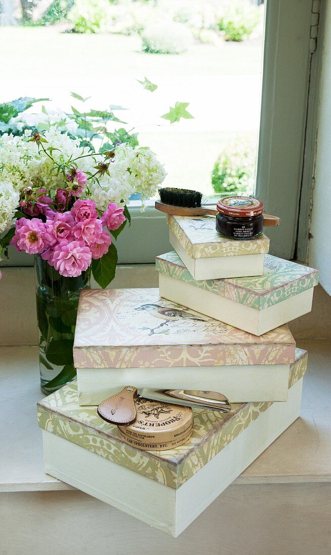 Stacked vintage boxes, shoe polish, shoe horn and vase of flowers on windowsill