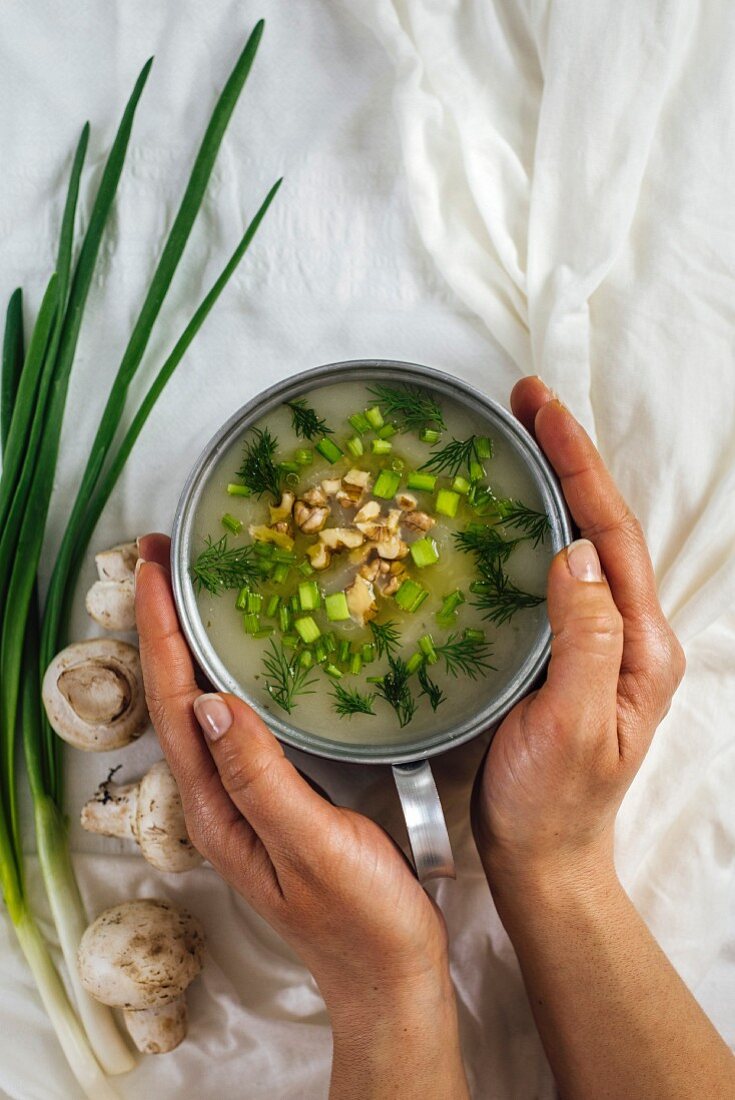 A woman is holding a bowl of cream of mushroom soup garnished with chopped walnuts and herbs