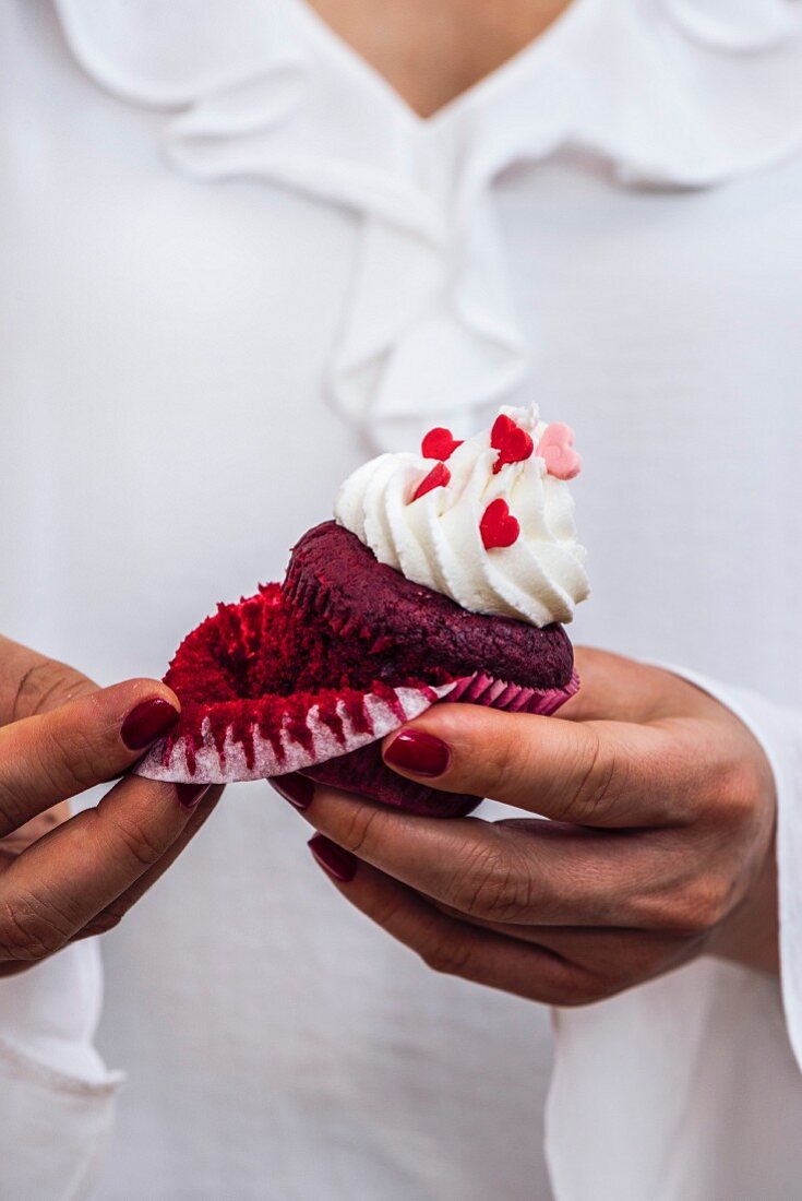 A woman with a white dress is holding a red velvet cupcake in her hand, removing the liner
