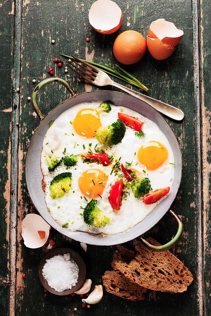 Pan of fried eggs, broccoli and cherry-tomatoes with bread on old green background