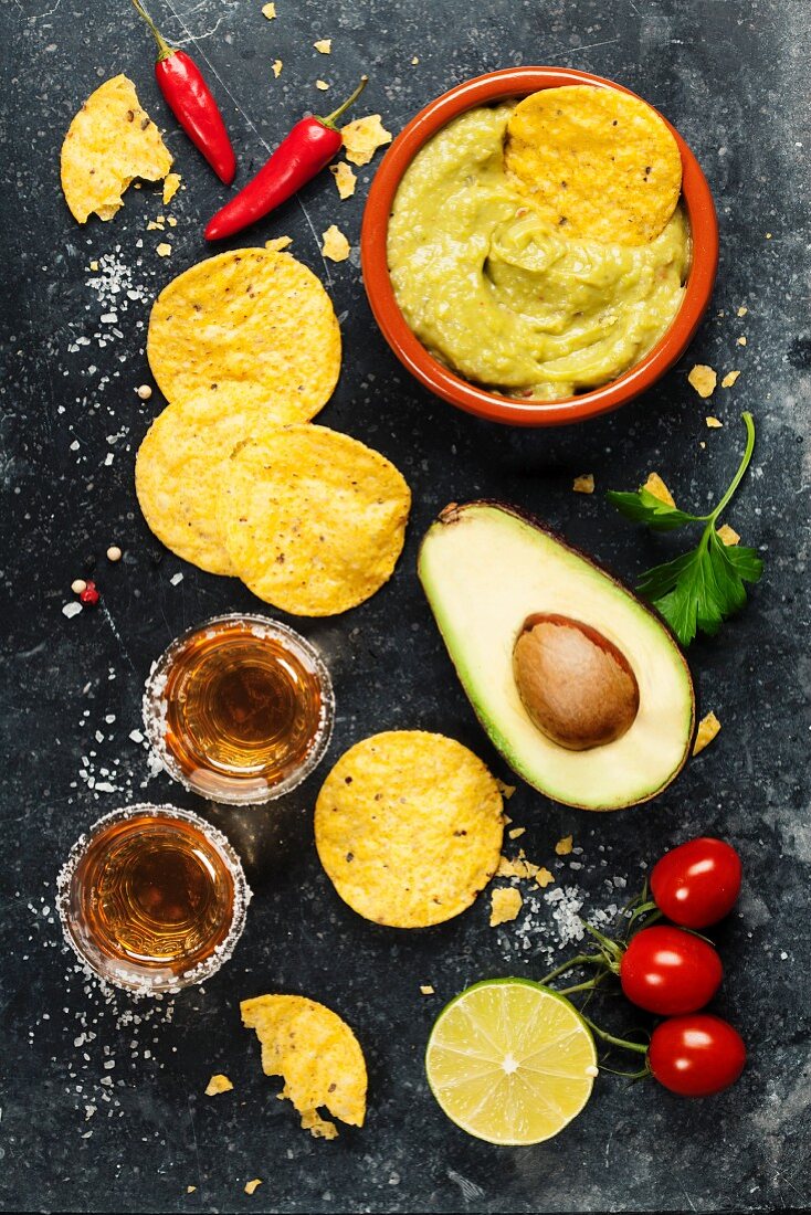 Bowl of mexican nachos chips with homemade fresh guacomole sauce and tequila shots over old background