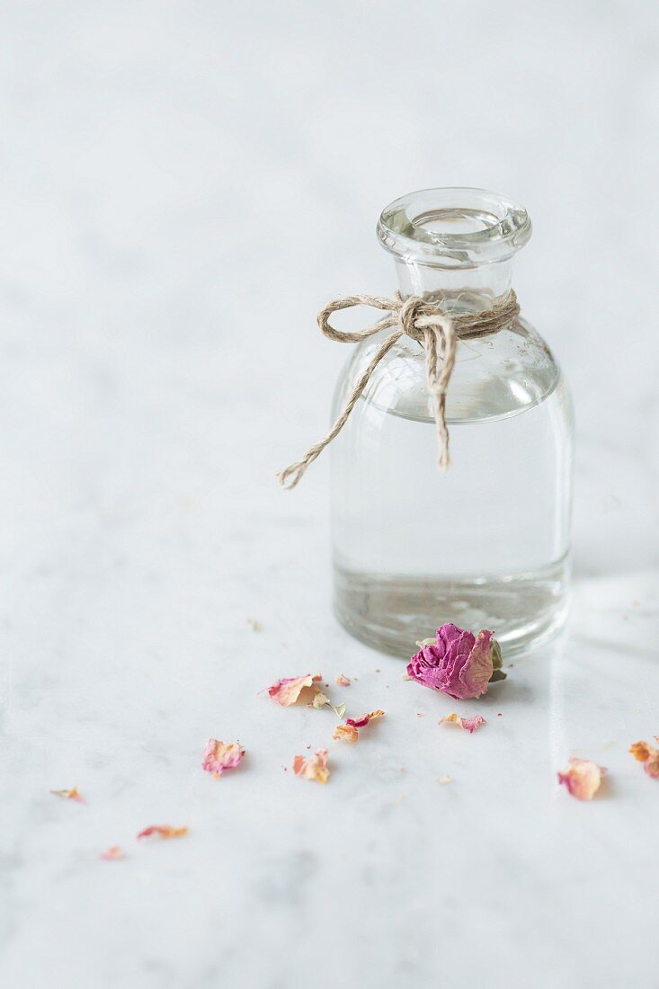 Clear glass bottle of rosewater tied with string on a white and grey marble surface with dried pink edible rose petals