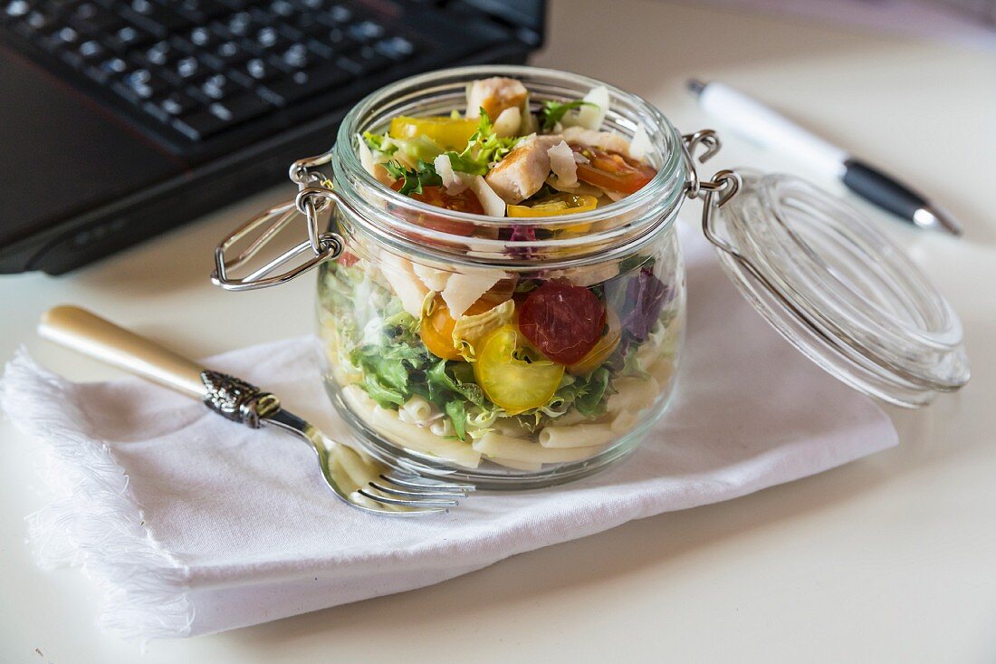 A pasta salad in a glass jar with tomatoes, chicken strips and pecorino