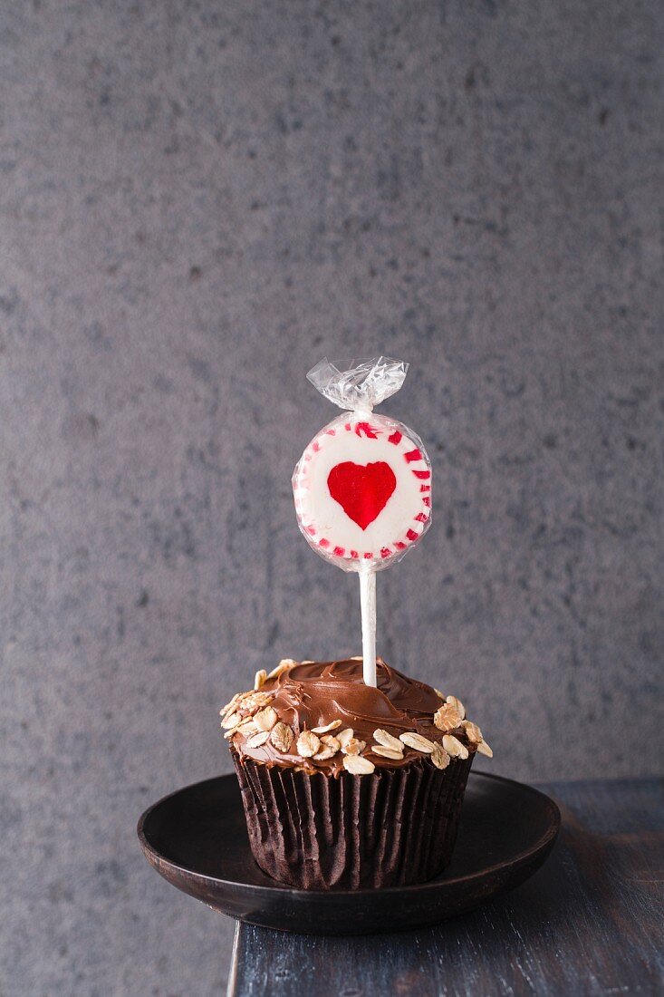 A cupcake with chocolate icing and oats with a heart-shaped lolly stuck in the top