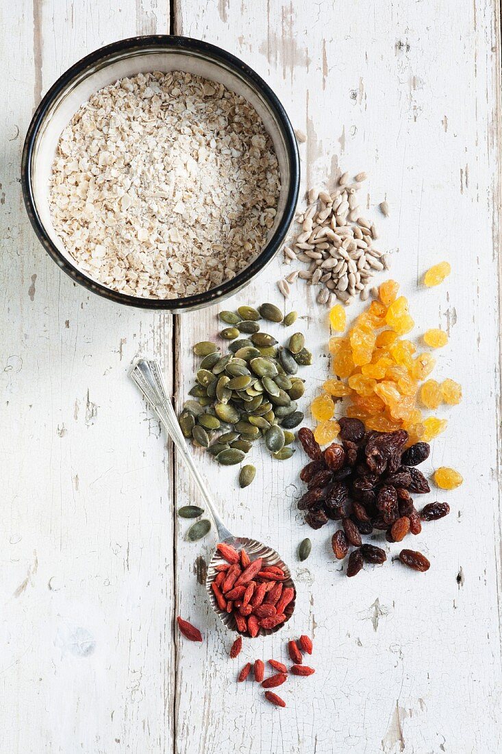Raisins, golden raisins, pumpkin seeds, sunflower seeds and goji berries in small piles, and a bowl of oats