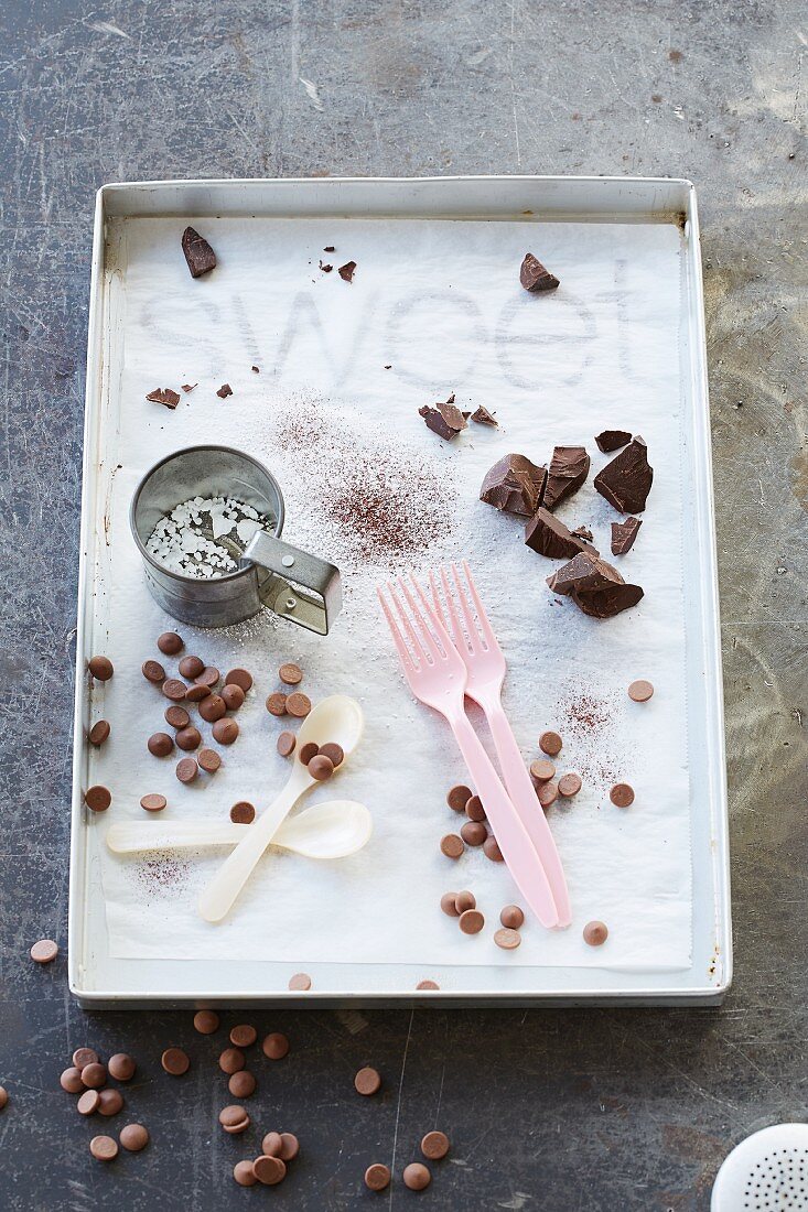 Chocolate drops, chocolate couverture, cocoa powder, a flour sieve, spoons and forks on a metal tray