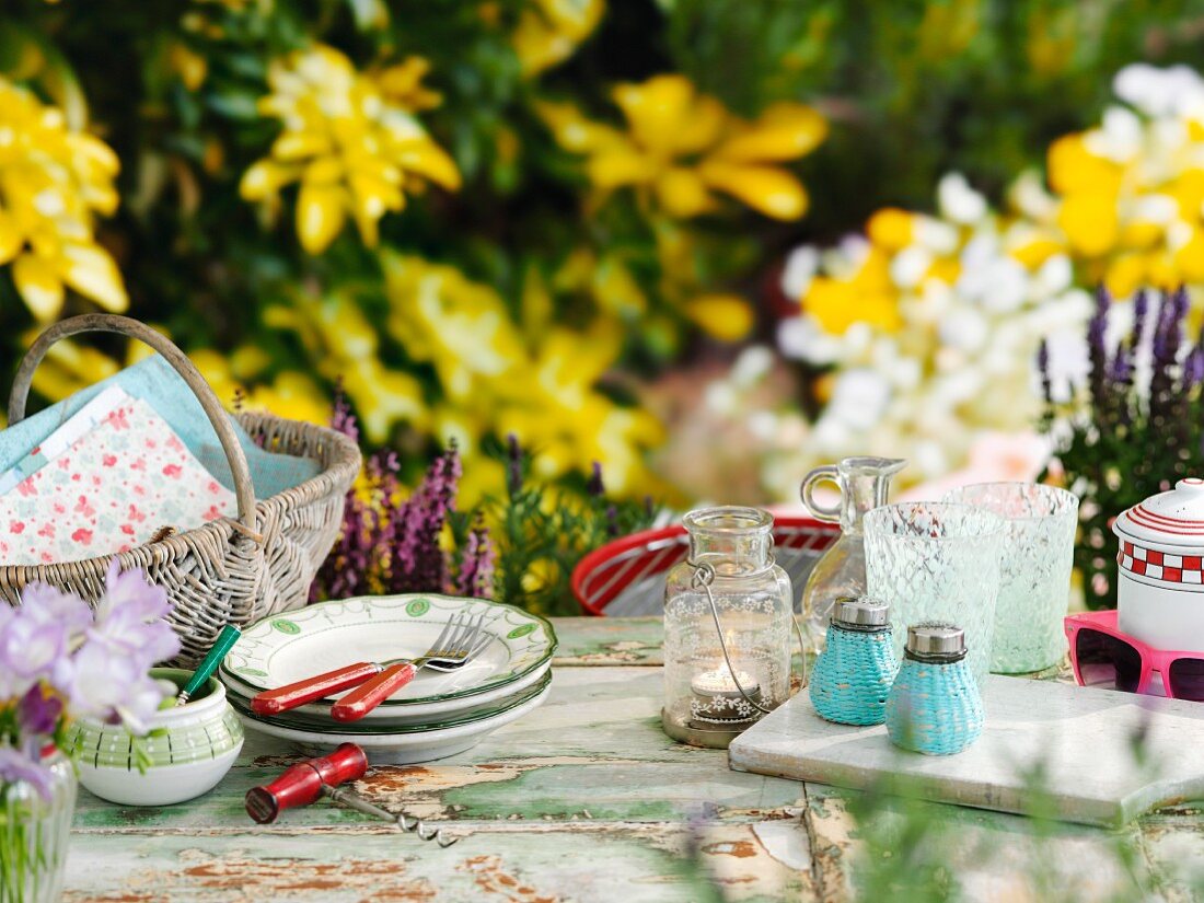 Korb, Geschirr, Windlicht, Gläser, Salz- und Pfefferstreuer auf Tisch im sommerlichen Garten