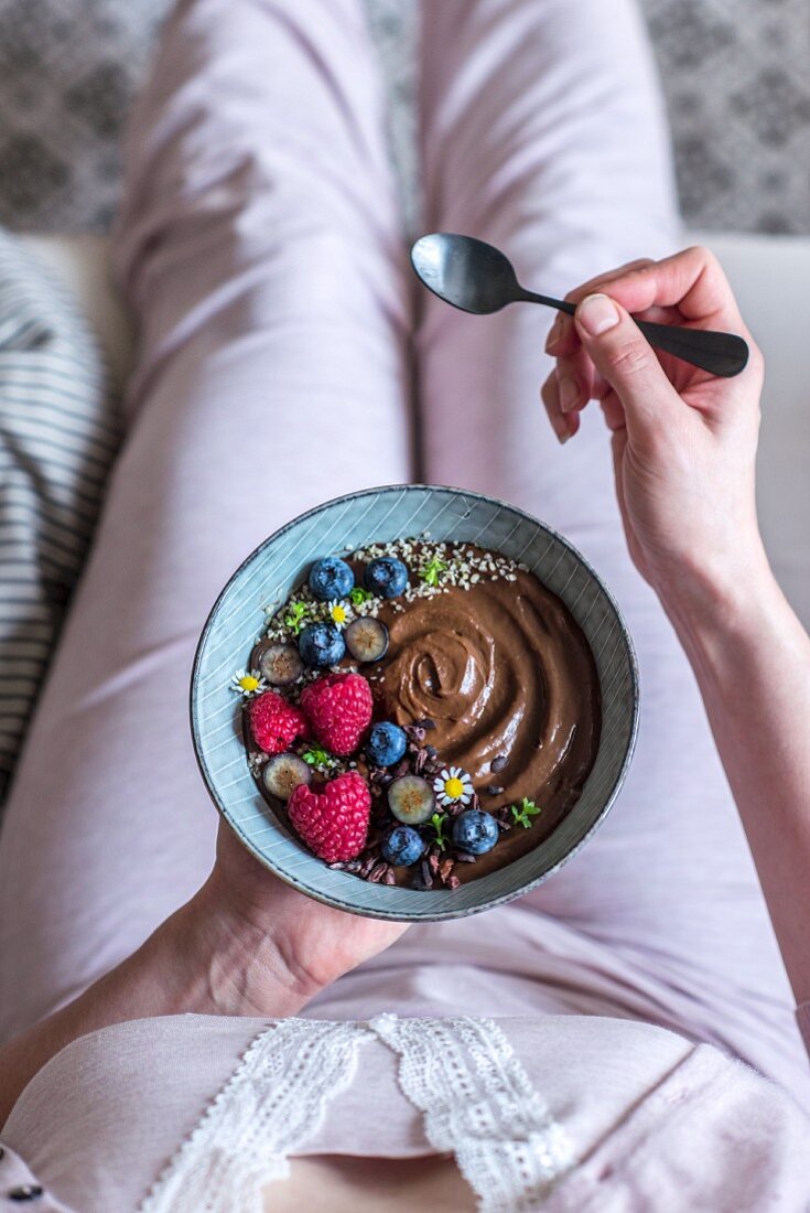 A woman holding a bowl of chocolate avocado mousse with banana, coconut oil, blueberries, strawberries, woodruff, chocolate chips and chamomile