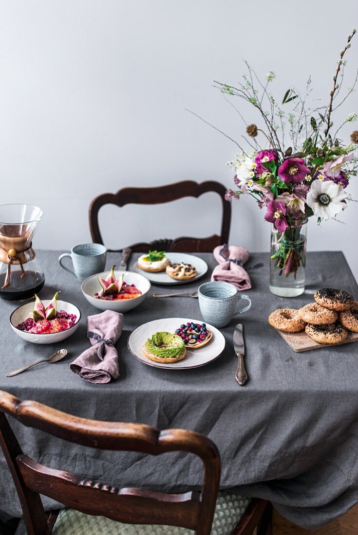 Frühstückstisch mit Hirse-Porridge, Dinkel-Bagels, Avocado, Blaubeeren, Granatapfel, Kakaonibs und Blumenstrauss