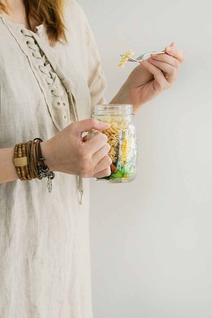 A woman in a linen dress holding a salad jar with wholegrain pasta, carrots, cauliflower and green salad leaves