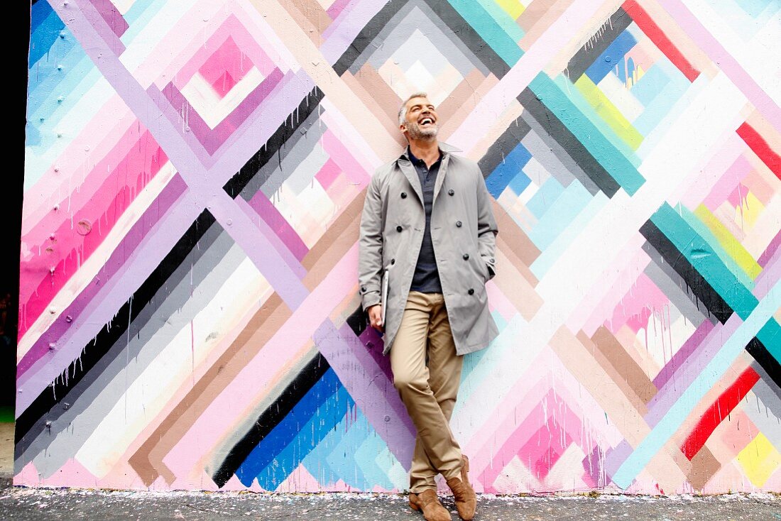 A man with grey hair wearing a coat and chinos standing in front of a brightly coloured wall