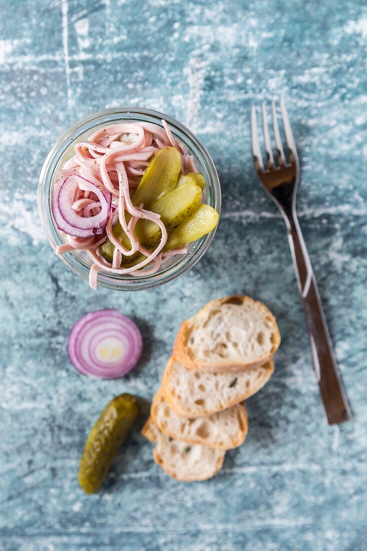 Wurstsalat mit roter Zwiebel und Essiggurken im Glas, daneben Baguettescheiben