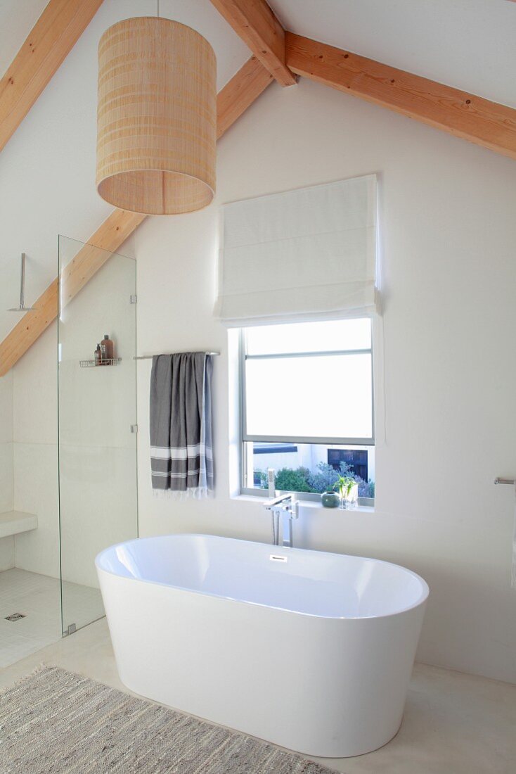 Free-standing white bathtub below window in gable wall in bright, modern bathroom