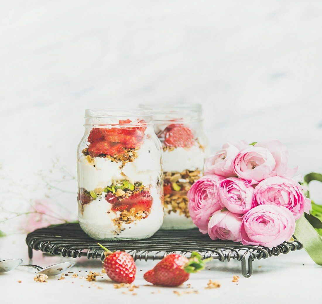 Greek yogurt, granola, strawberry breakfast in glass jars, pink raninkulus flowers, marble background