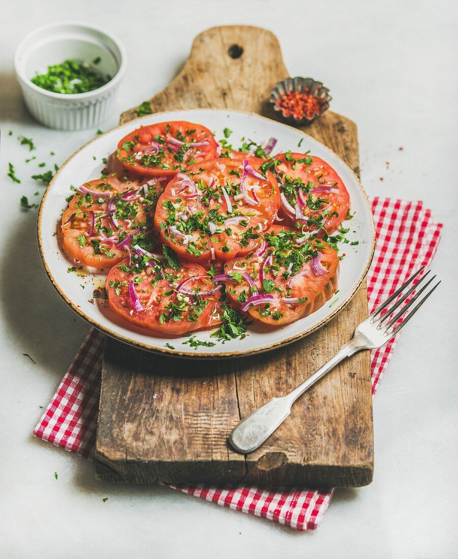 Fresh heirloom tomato, parsley and onion salad in white round plate on wooden board over light grey marble background