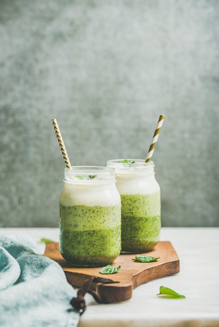 Ombre layered green smoothies with mint in glass jars with straws on wooden board, grey concrete wall background