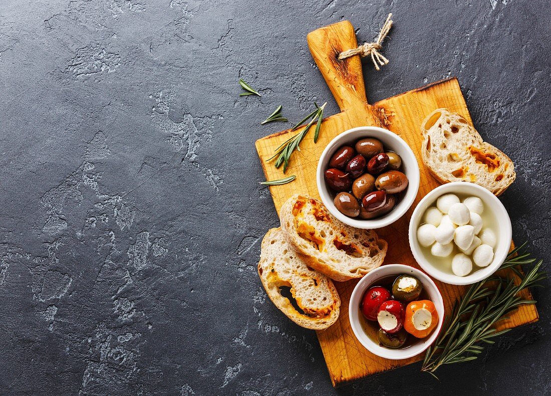 Antipasti: Oliven, gefüllte Paprika, Mini-Mozzarella und Ciabatta auf Holzbrett (Aufsicht)