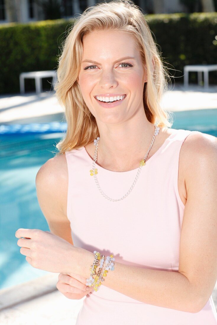 A blonde woman wearing a pink dress and a necklace in front of a pool