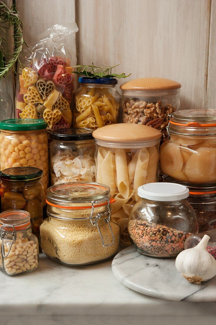 Vorratsgläser mit Nudeln, Nüssen, Marmelade, Oliven, Obst und Linsen auf Marmorplatte
