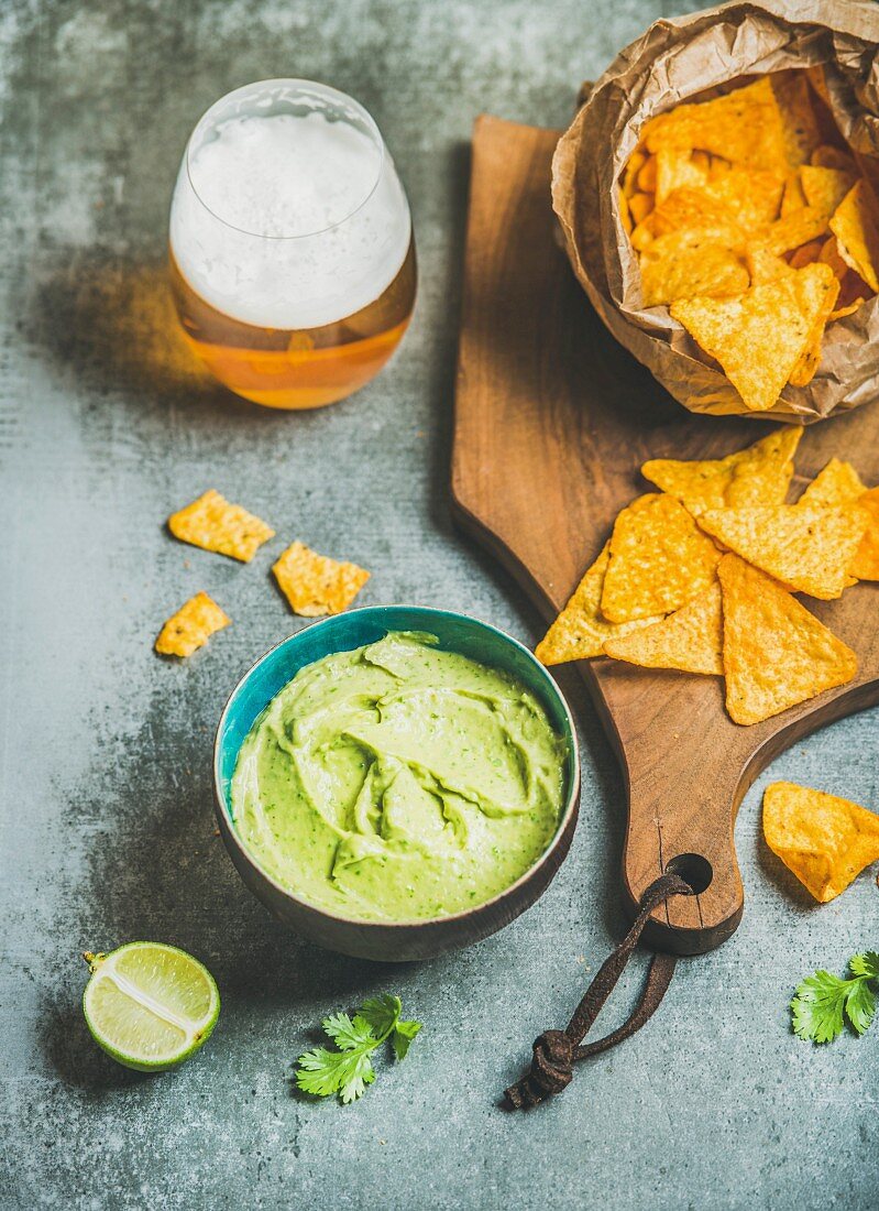 Mexican corn chips, fresh guacamole sauce and glass of beer on wooden serving board over grey concrete table background, selective focus, vertical composition