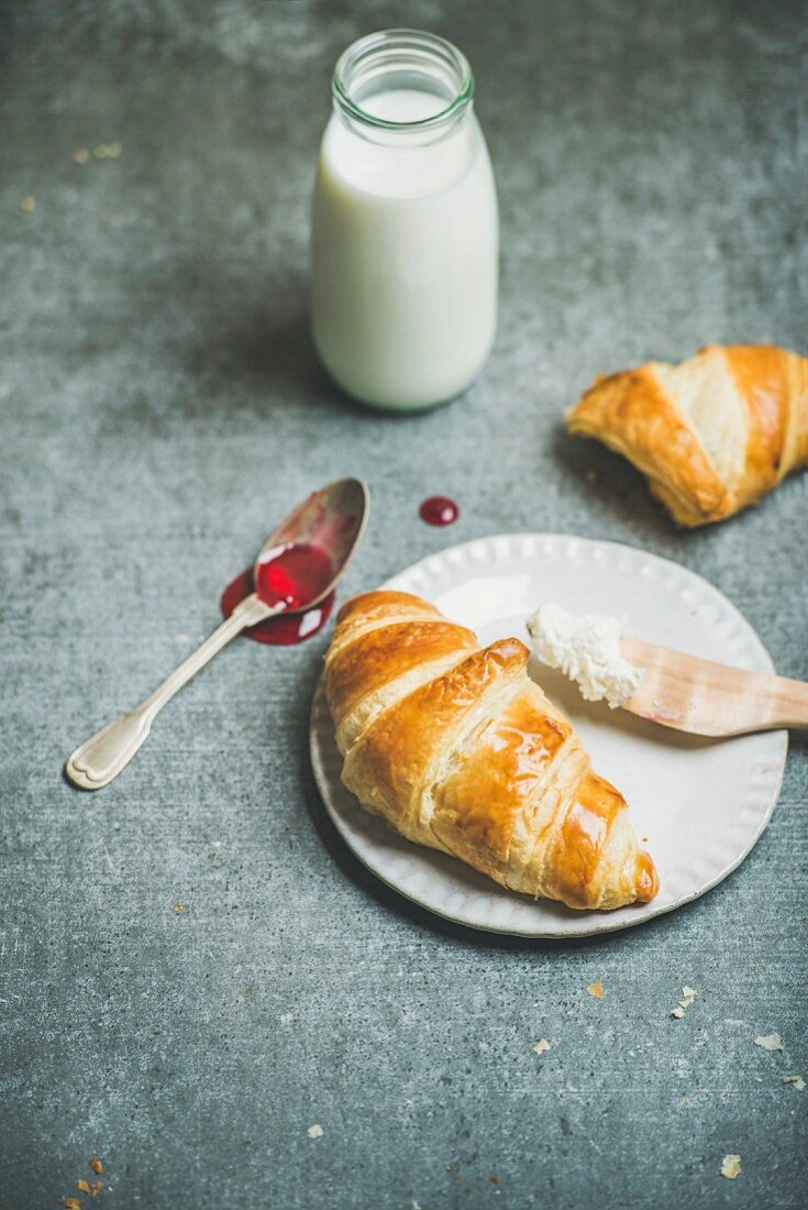 Freshly baked croissants with raspberry jam, ricotta cheese and milk in bottle over grey concrete background