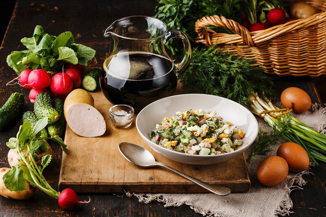 Cold Soup Okroshka with sausage, vegetables and kvass serving size in bowl and raw ingredients on wooden table