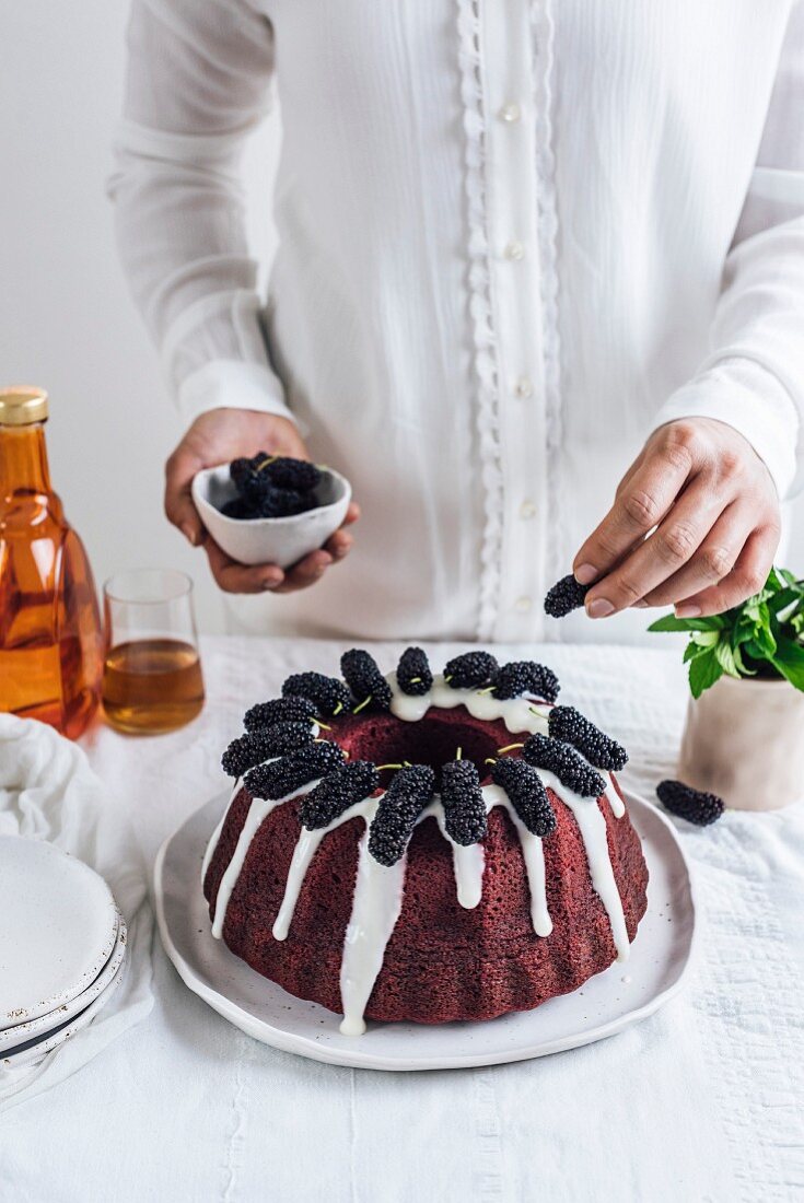 A woman with white shirt topping a red velvet bundt cake with black mulberries