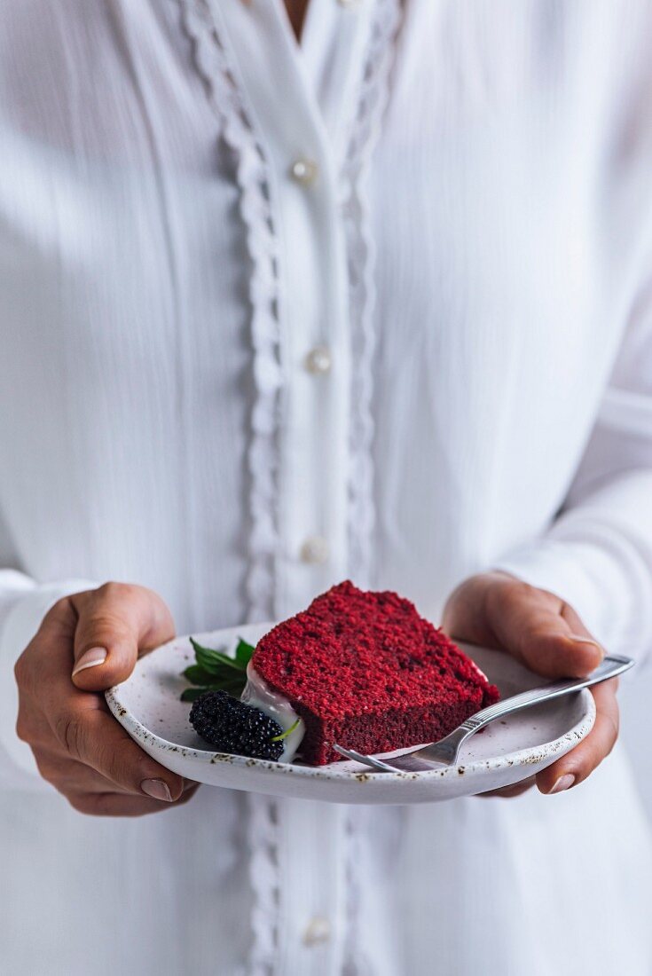 A woman with white shirt holding a slice of red velvet bundt cake with white glaze, a mulberry and mint
