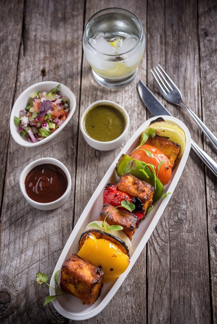 A paneer and vegetable skewer with different sauces