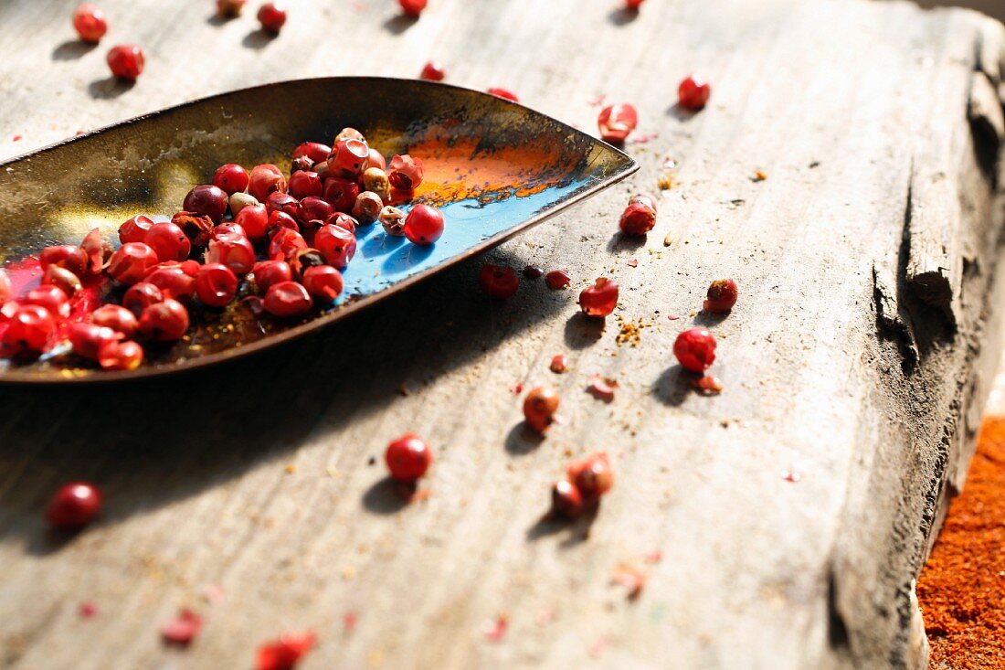 Pink pepper in and next to a small metal scoop on a wooden surface