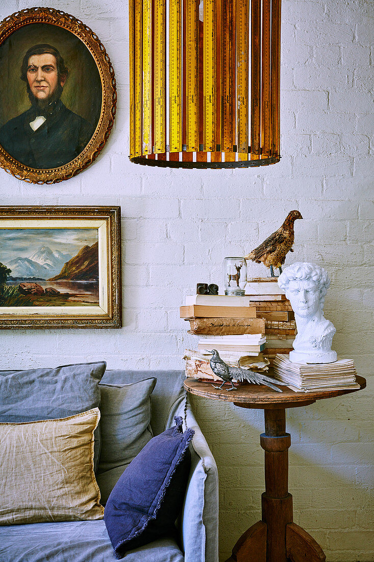 Antik Holztisch mit Büste, Büchern und Vogelfiguren neben Sofa im Wohnzimmer mit weiss gestrichener Ziegelwand
