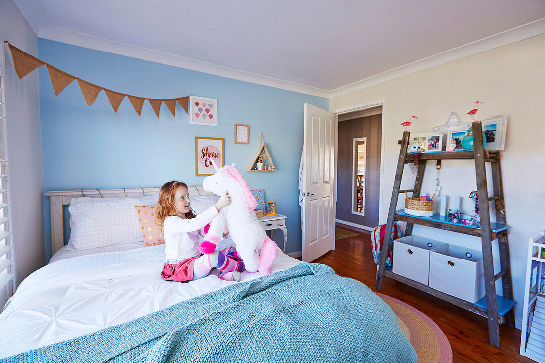 Girl plays on bed, DIY shelf made of old wooden ladder in children's room with light blue wall