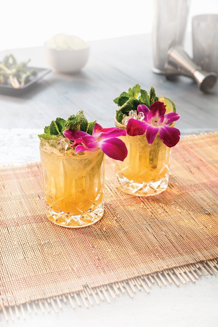 Two Mai Tai cocktails garnished with orchid blossoms and mint in crystal glasses