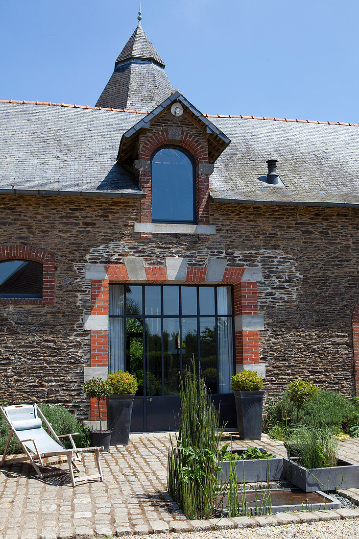 Terrasse mit Teich und Pflanzkübeln vor altem Steinhaus mit Glastür