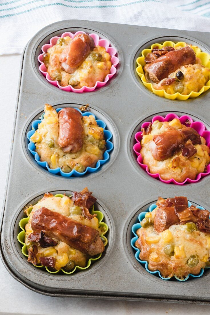 A metal muffin tray with savoury Frankfurter sausage muffins in colourful silicone cases. Crispy bacon, corn and green peas