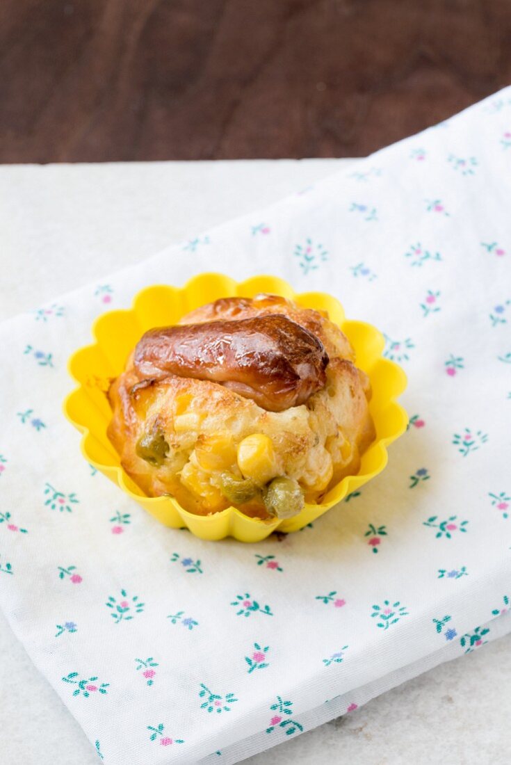 A savoury muffin with a Frankfurter sausage, corn and peas in a yellow muffin case on top of a floral napkin
