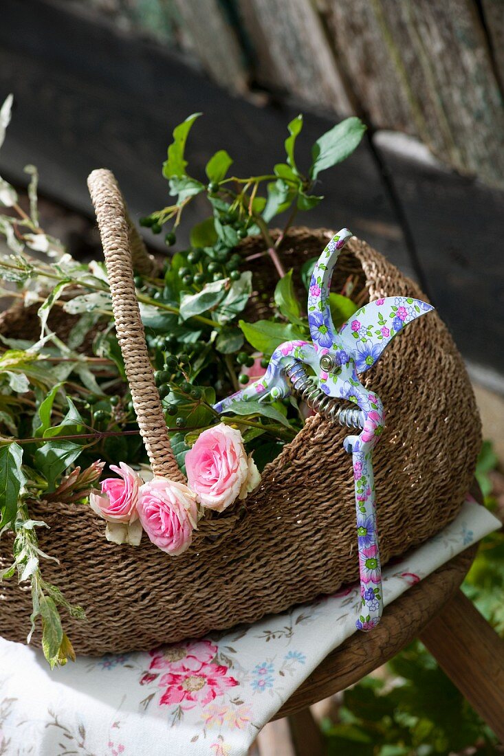 Henkelkorb mit Rosenblüten, grünen Zweigen und Gartenschere