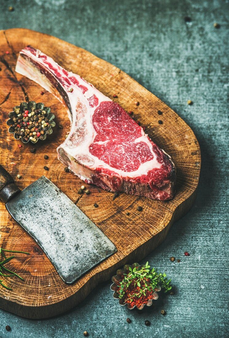 Dry aged raw beef rib eye steak with bone, butcher meat chopping knife and spices on rustic wooden board