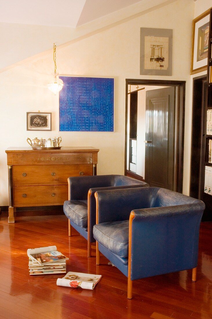 Two blue leather armchairs in front of old chest of drawers and blue picture