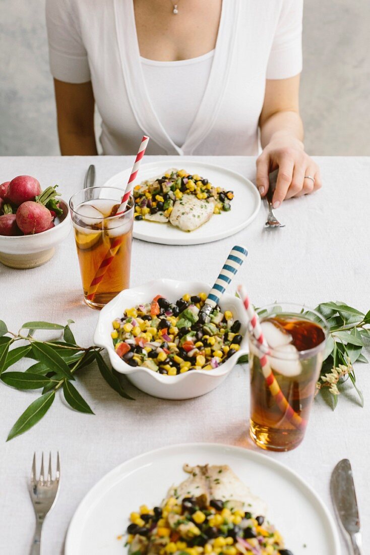 A woman is photographed at a tablesetting with tilapia and black bean salsa in her plate