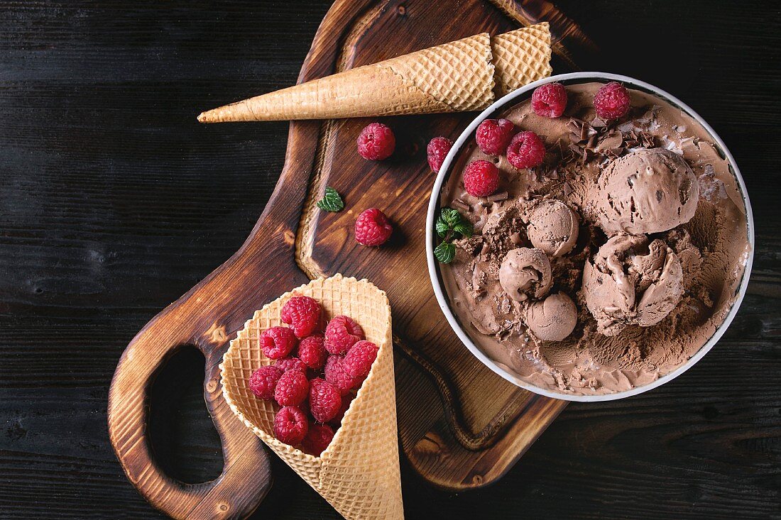 Bowl of homemade chocolate ice cream with fresh raspberries, mint and waffle cone on wood serving board