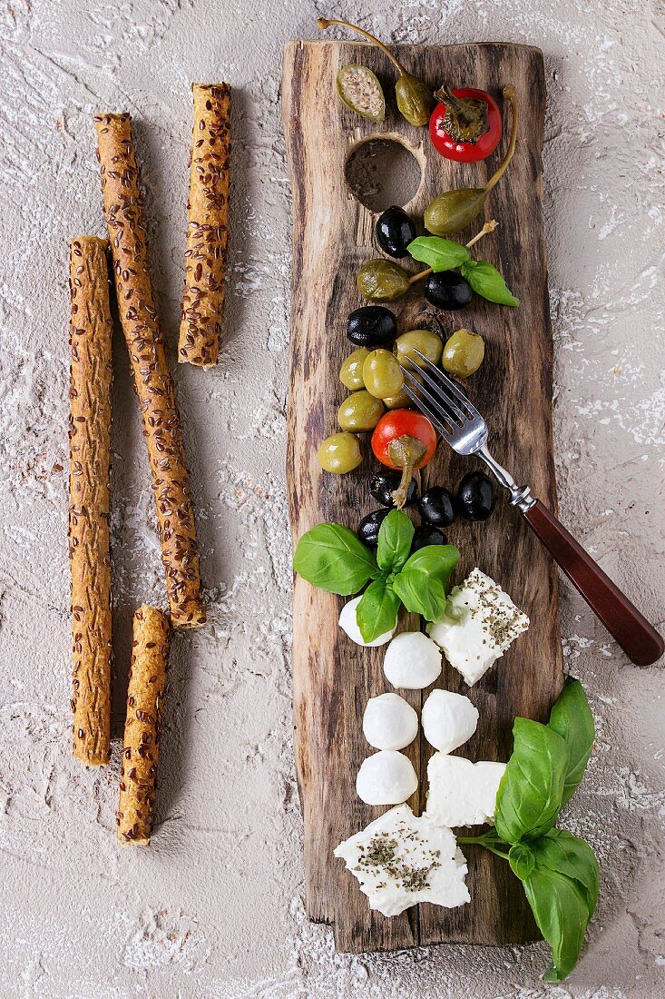 Grissini mit Oliven, Kapern, Feta und Mozzarella als mediterrane Antipasti auf Holzbrett