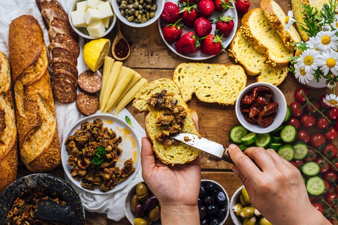 A woman spreading olive tapenade on a slice of corn bread, French baguette, sausages, baby corns, cheese, strawberries
